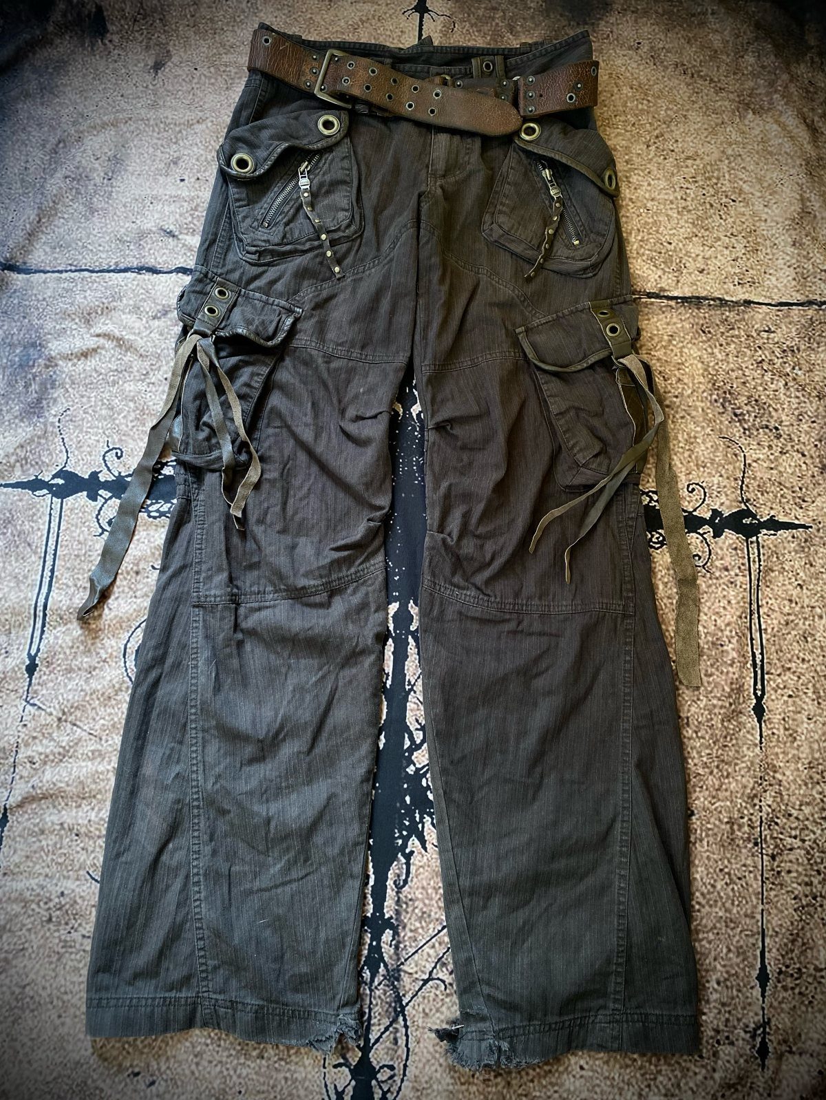 Dark olive green cargo pants with utility pockets and straps laid on a textured surface.