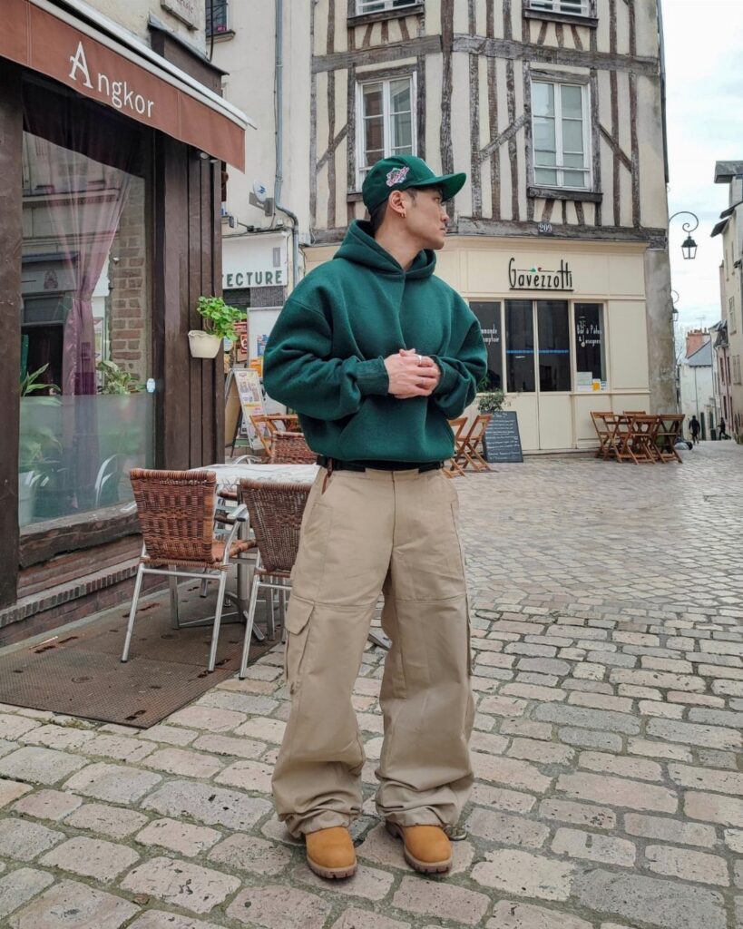 A man in an oversized green hoodie and baggy tan cargo pants standing on a cobblestone street.